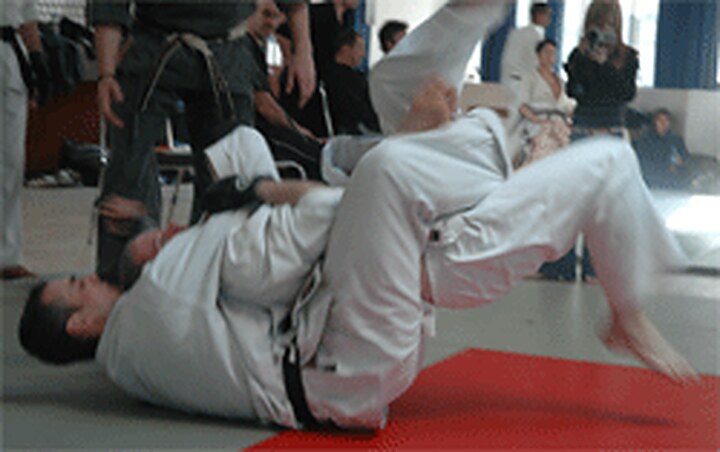 A throw in progress on the red mat, one karateka on their back sending a partner over the top, watched by a hall of students